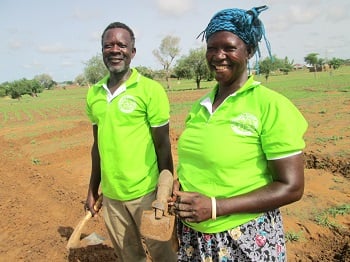 Mr. Akolgo, left, with close cropped hair and grey beard, stands next to Madam Adukpoka on the right. They both wear lime green shirts and are standing in a recently tilled field, holding farm tools.