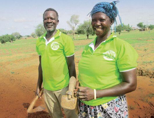 Akolgo and Adukpoka stand in front of the demonstration farm smiling with basic farming tools in their hands.