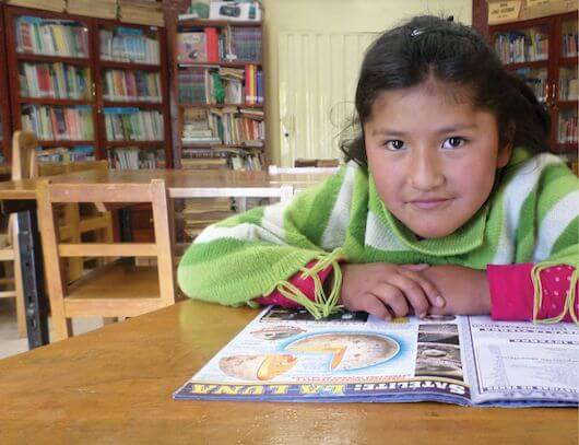 Eight-year-old Araceli sits at a desk reading a magazine in a classroom filled with desks, chairs and a bookcase well-stocked with books.