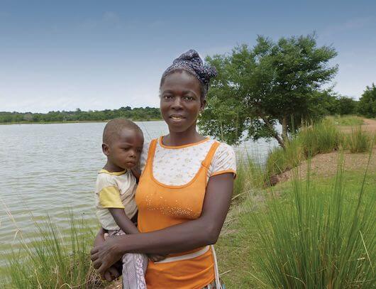 Christina stands on the banks of a river holding her young child in her arms.