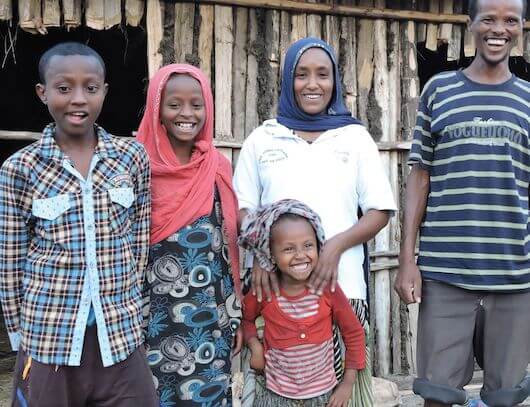 Hawa stands behind her youngest child with two of her children (a boy and a girl) standing off to her right. Her husband stands to her left. They are standing outside their wooden home laughing and smiling.