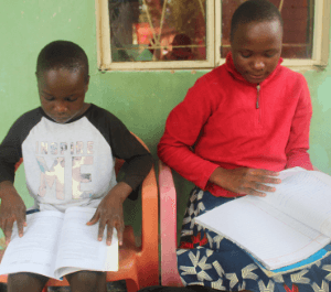 Two young children sit in chairs in front of their home studying