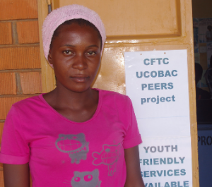 Teenage girl stands next to sign that says "CFTC & UCOBAC Peers Project. Youth Friendly Services Available"