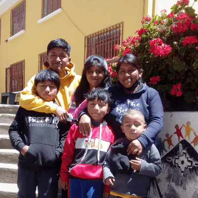 Lidia in the top right, smiles with four of her children, including only daughter Priscilla to her right. They are outdoors in front of an flowering bush and some steps. 